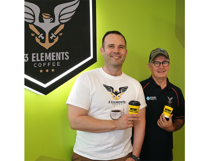 Two men standing in front of a coffee shop with a logo, showcasing a vibrant and inviting atmosphere.