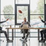 smiling young businessmen using laptops and playing with soccer and rugby balls in office