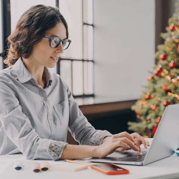 Businesswoman wearing glasses sitting in office near xmas tree and working on laptop during the holiday season