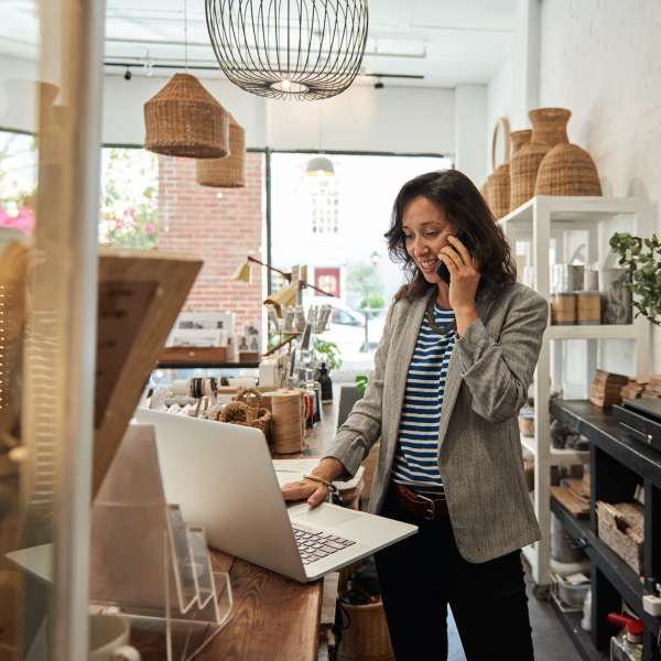 Smiling young woman standing behind a counter in her stylish boutique working on a laptop and talking on a cellphone