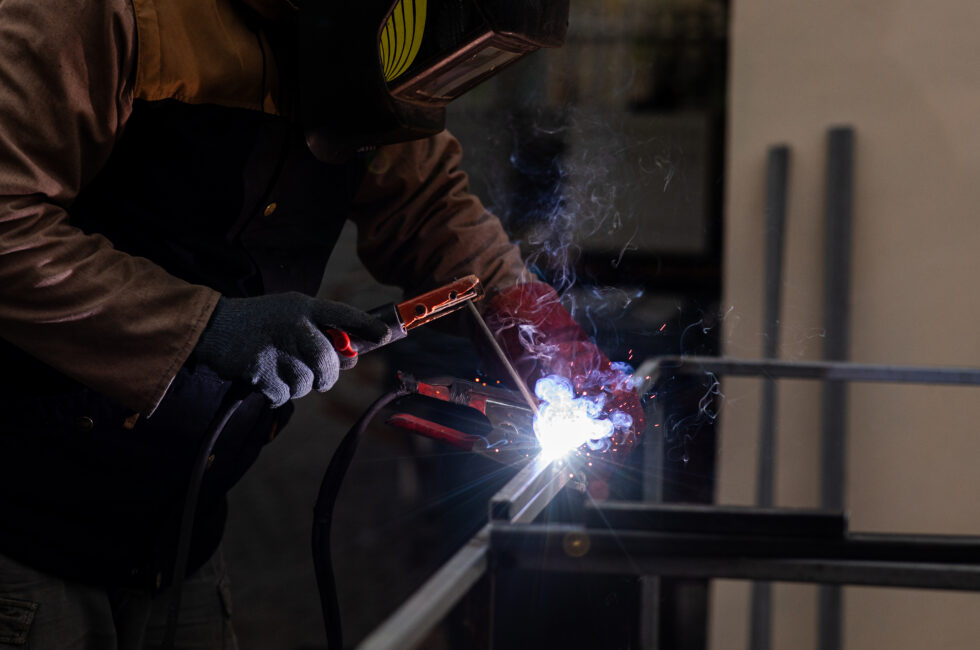 A worker is welding a metal frame in a workshop. The scene shows bright sparks and smoke around the welder.
