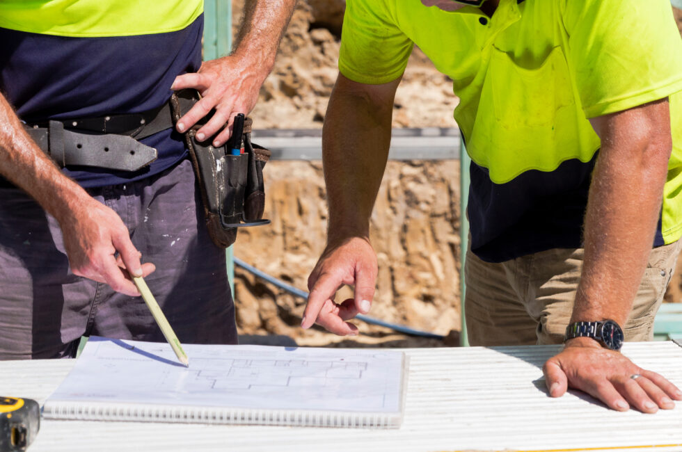 Two builders on site look at house plans and discuss.