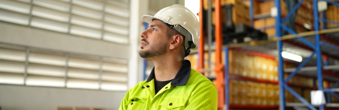 A manager in a hard hat and fluros standing alone in a warehouse.