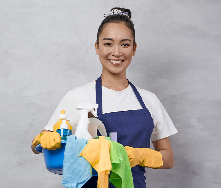 A woman in an apron holds a bucket filled with various cleaning supplies
